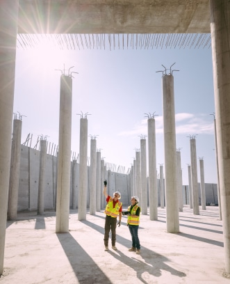 Dos trabajadores con equipo de seguridad observan una obra en construcción con grandes postes y el cielo de fondo.